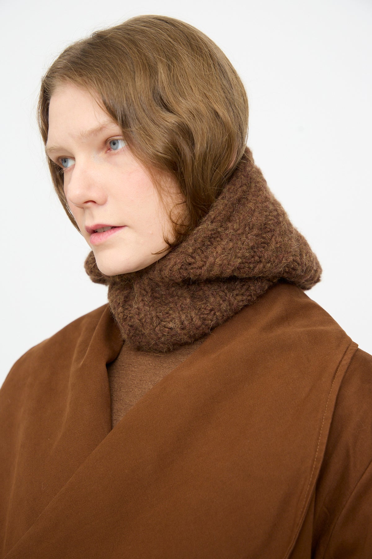 A person with chin-length brown hair wearing a brown coat and the Lauren Manoogian Handknit Rib Balaclava in Rust Melange, looking to the left against a plain light background.