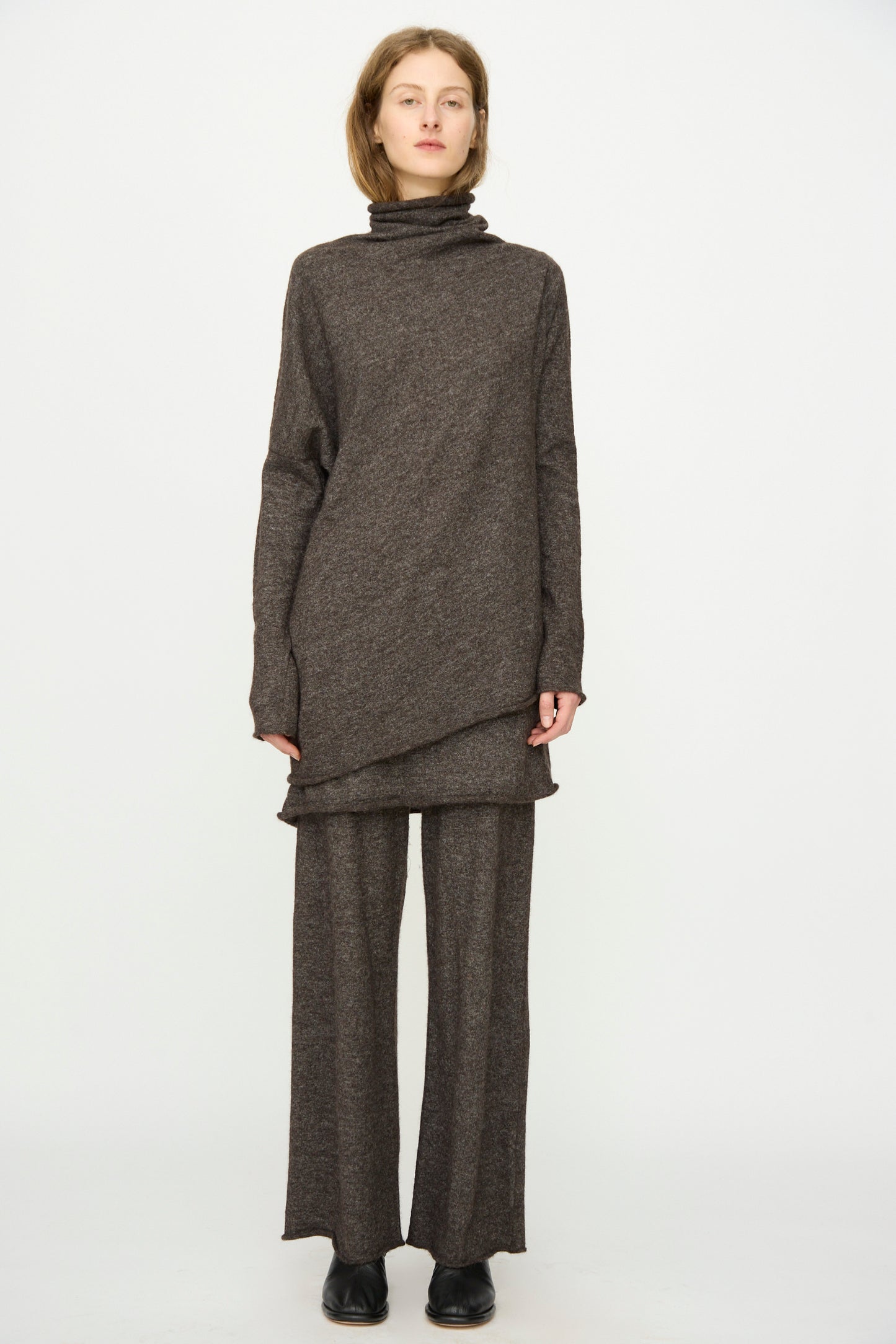 A person stands facing forward, wearing the Lauren Manoogian Askew Funnel Sweater in Blackened with relaxed wide-leg pants and black shoes, posing against a plain white background.