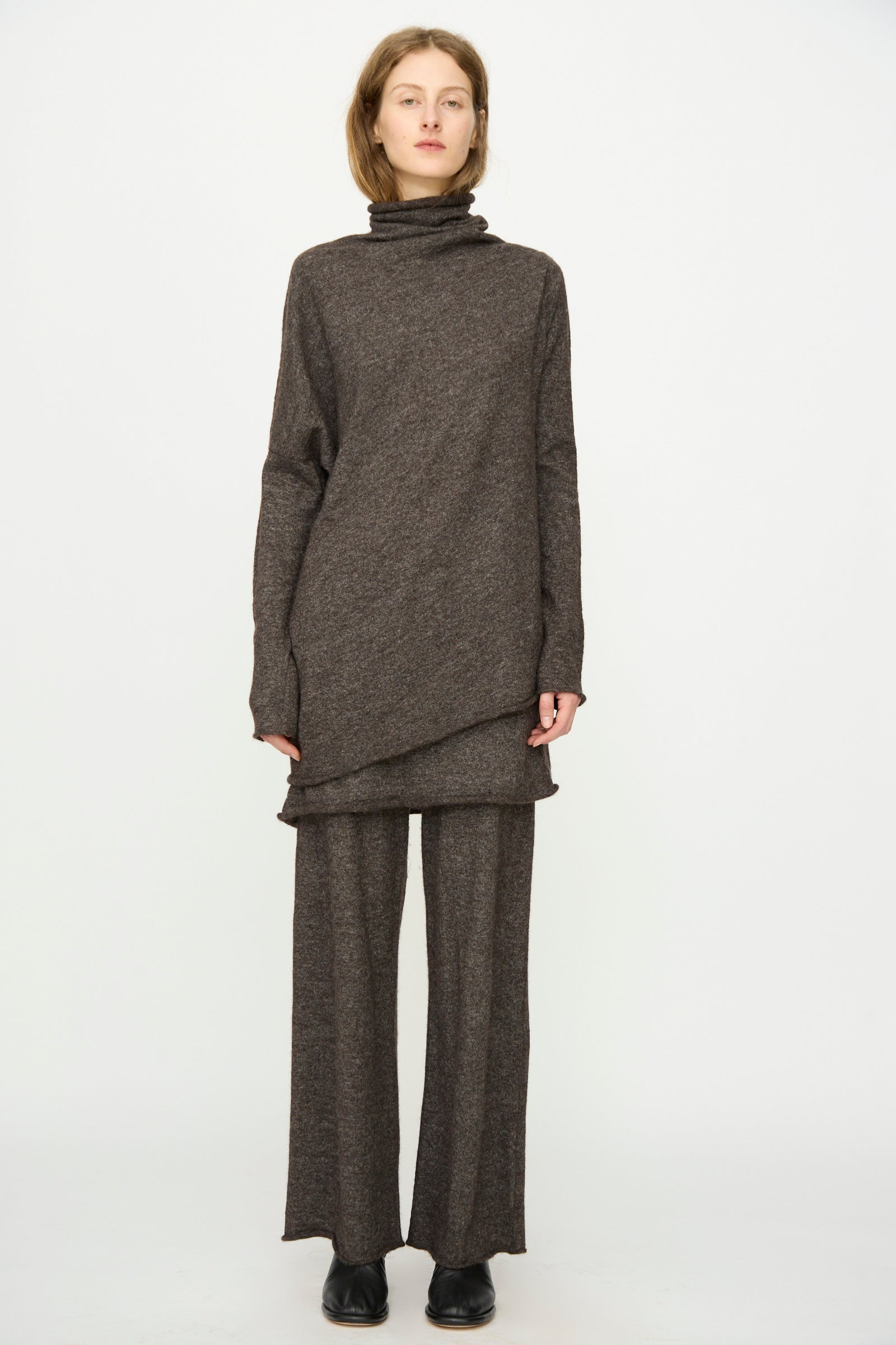 A person stands facing forward, wearing the Lauren Manoogian Askew Funnel Sweater in Blackened with relaxed wide-leg pants and black shoes, posing against a plain white background.