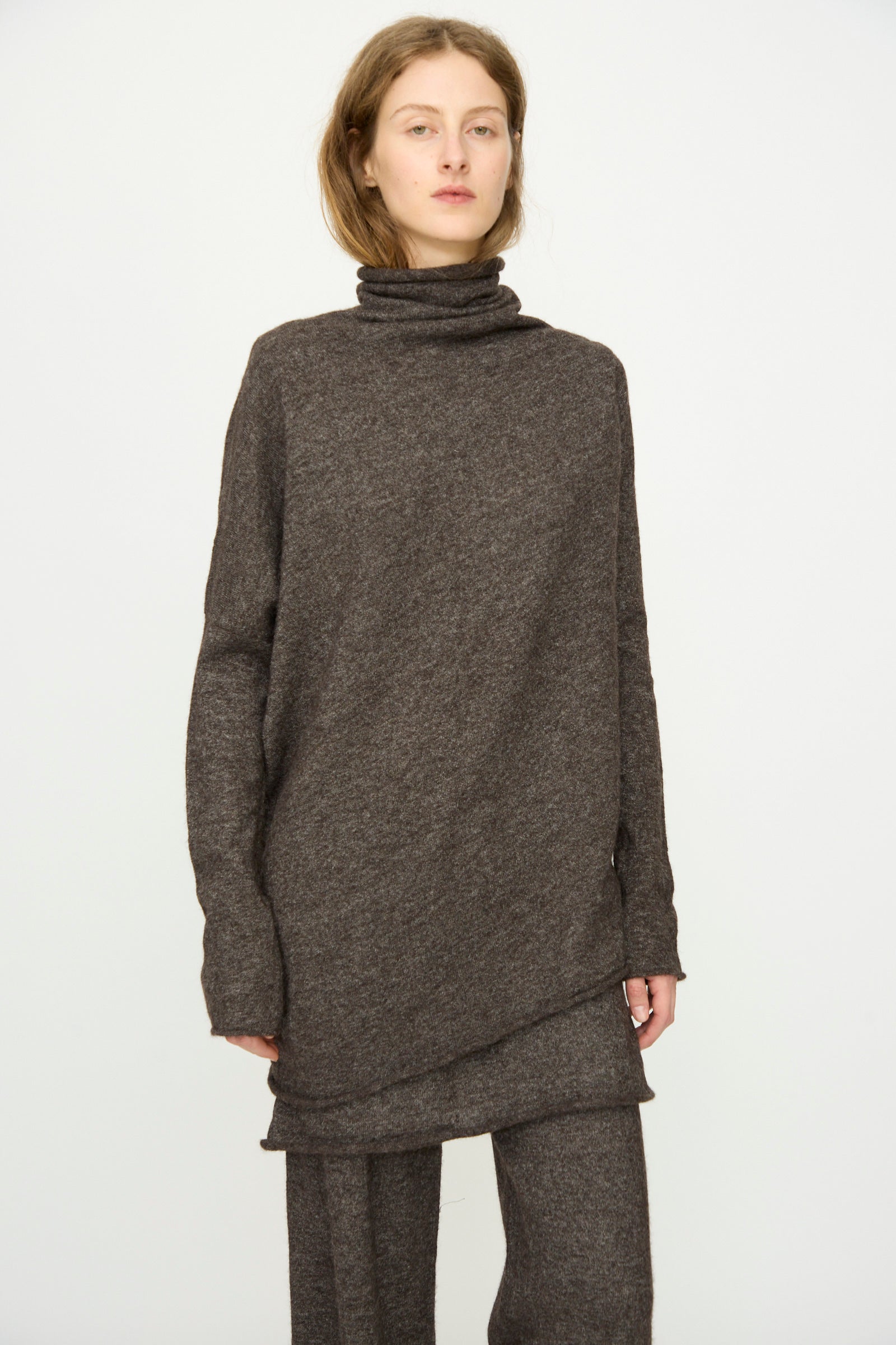 A person wears Lauren Manoogian's Askew Funnel Sweater in Blackened, a relaxed-fit, long-sleeved turtleneck, paired with matching pants, standing against a plain white background.