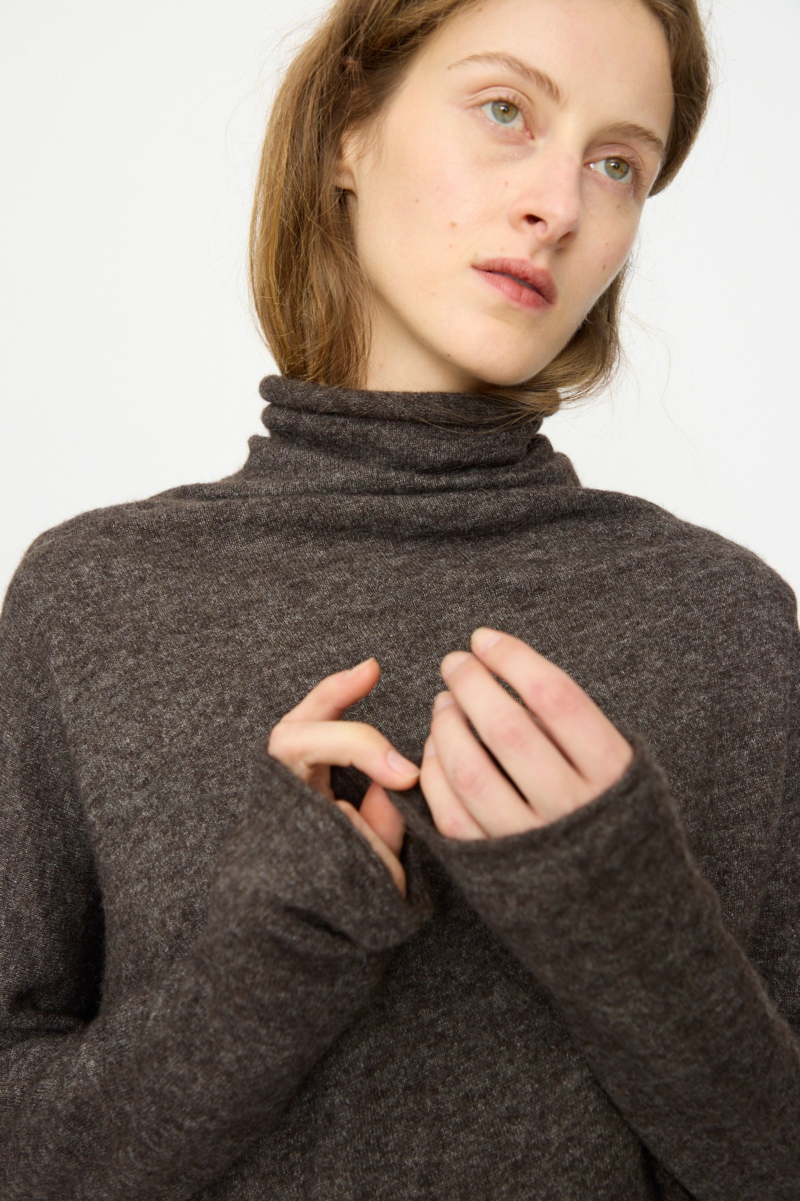 A woman models the Lauren Manoogian Askew Funnel Sweater in Blackened, standing against a plain background and gazing to the side with her hands raised near her chest.
