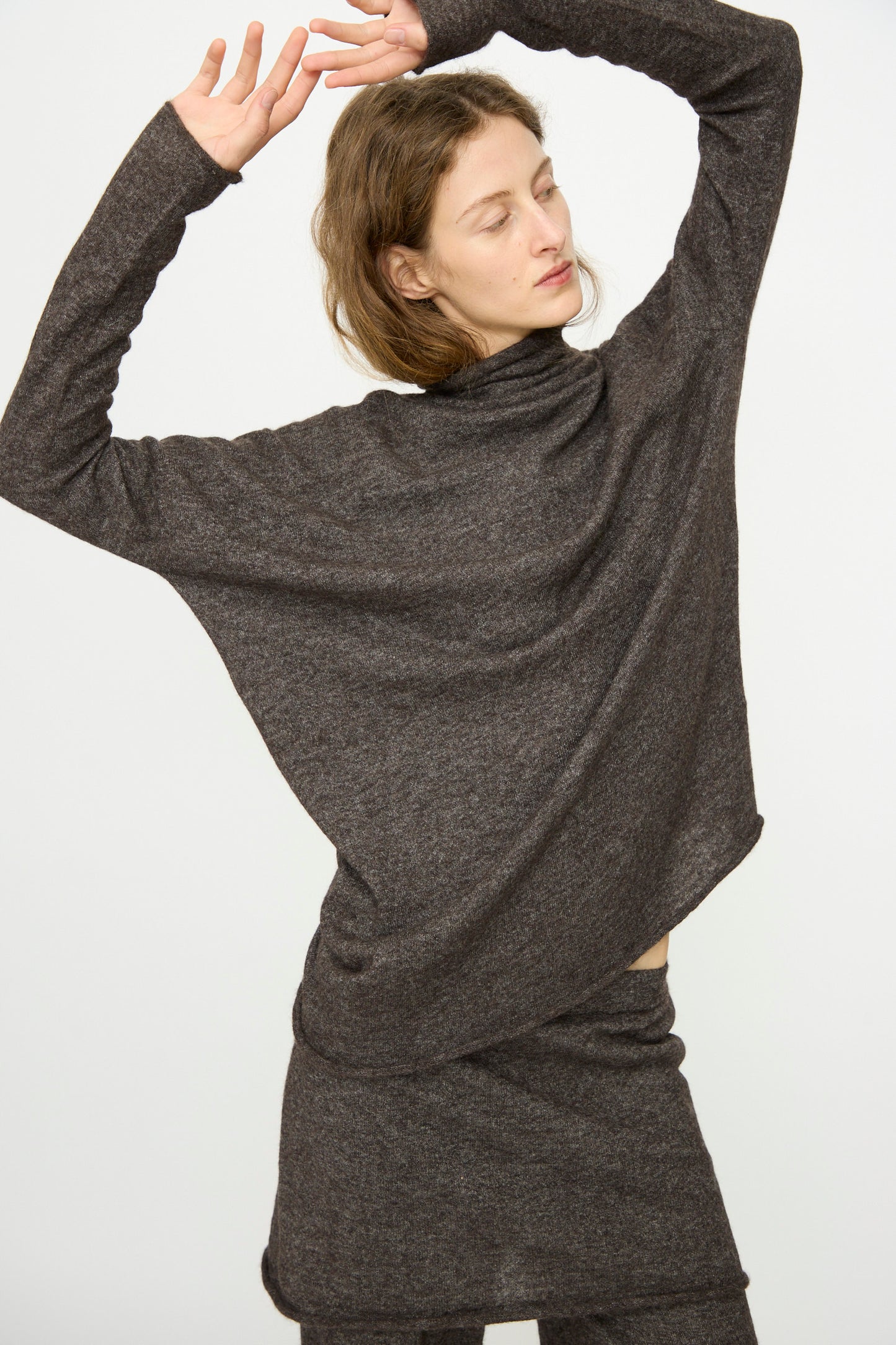 A woman poses with arms raised, wearing the Lauren Manoogian Askew Funnel Sweater in Blackened, styled with a matching skirt, against a plain white background.