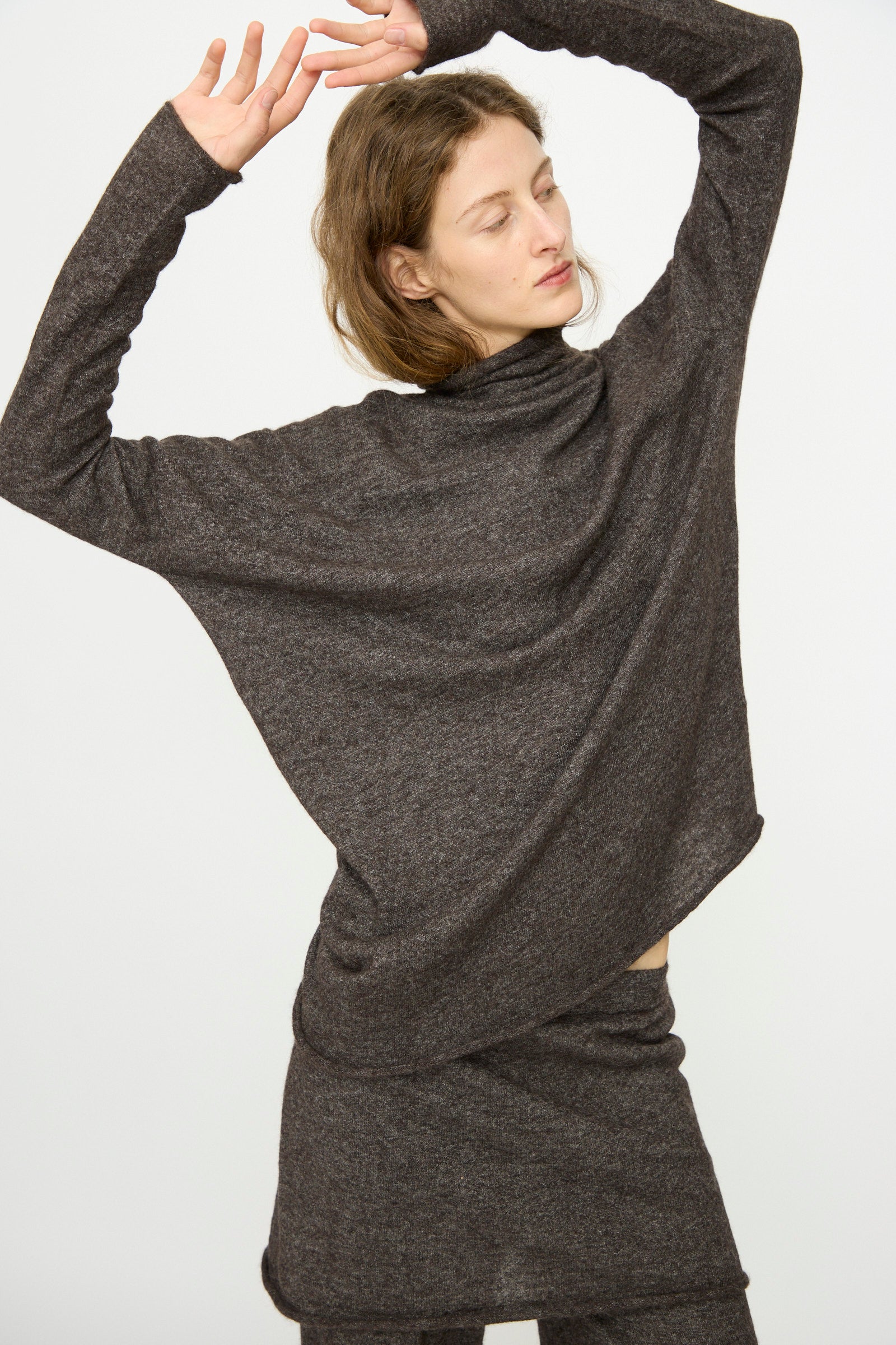 A woman poses with arms raised, wearing the Lauren Manoogian Askew Funnel Sweater in Blackened, styled with a matching skirt, against a plain white background.