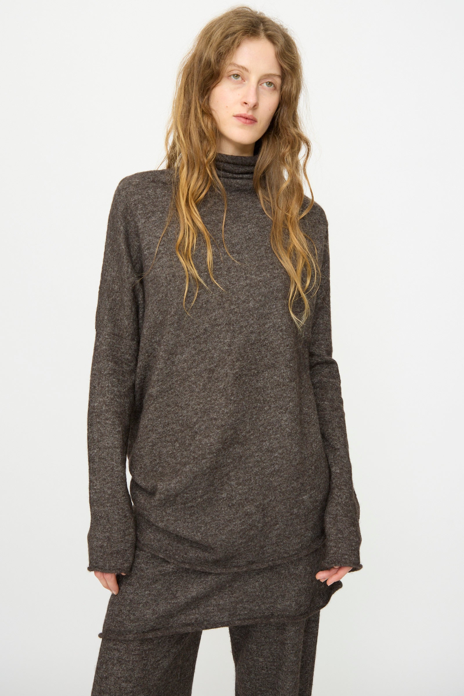A person with long, wavy hair wears the Lauren Manoogian Askew Funnel Sweater in Blackened and matching pants, standing against a plain white background.