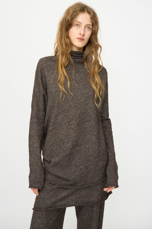 A person with long, wavy hair wears the Lauren Manoogian Askew Funnel Sweater in Blackened and matching pants, standing against a plain white background.
