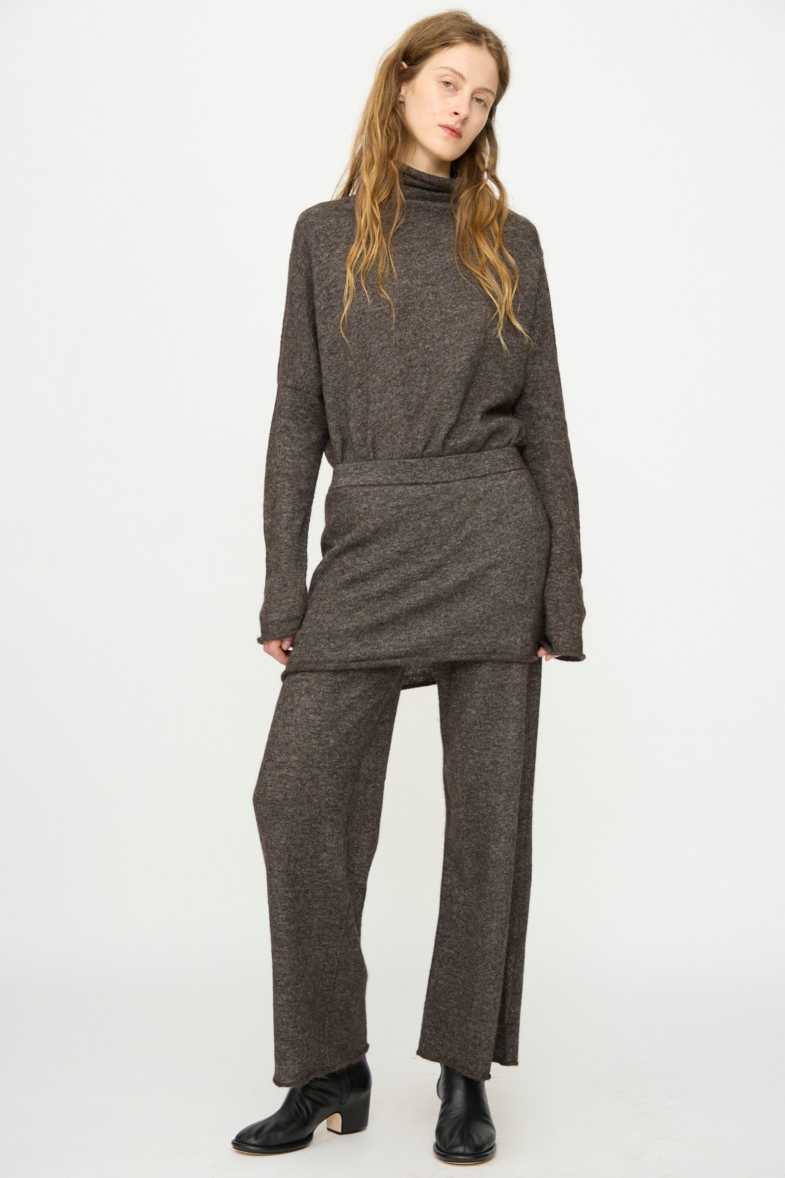 A woman stands against a plain background wearing Lauren Manoogian's Askew Layer Pants in Blackened, paired with a loose brown turtleneck tunic and black ankle boots.