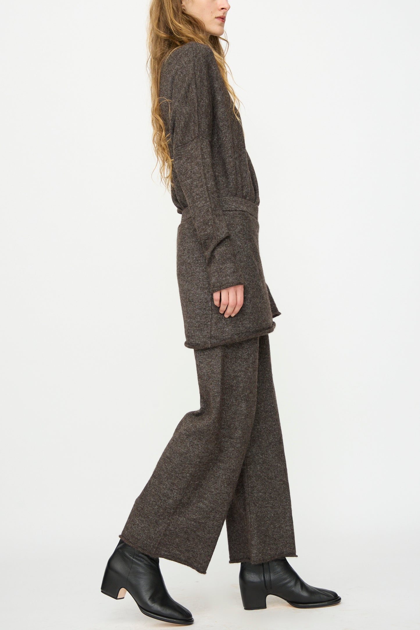 A person stands sideways in Lauren Manoogian Askew Layer Pants in Blackened, styled with a loose brown knit sweater and black heeled ankle boots, against a plain white background.