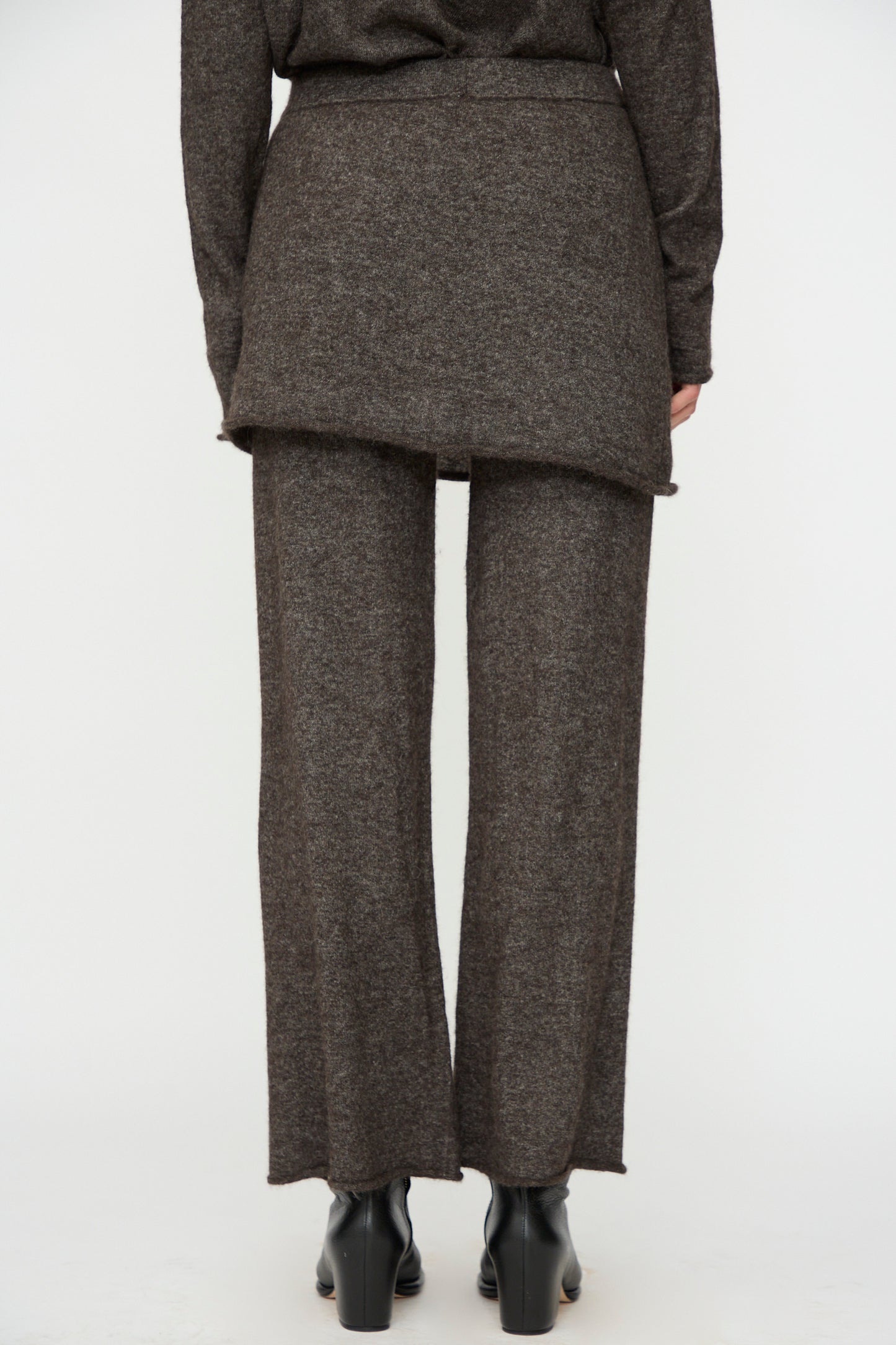 Person facing away, wearing Lauren Manoogian Askew Layer Pants in Blackened with a matching long top and black heeled shoes, standing against a plain light background.