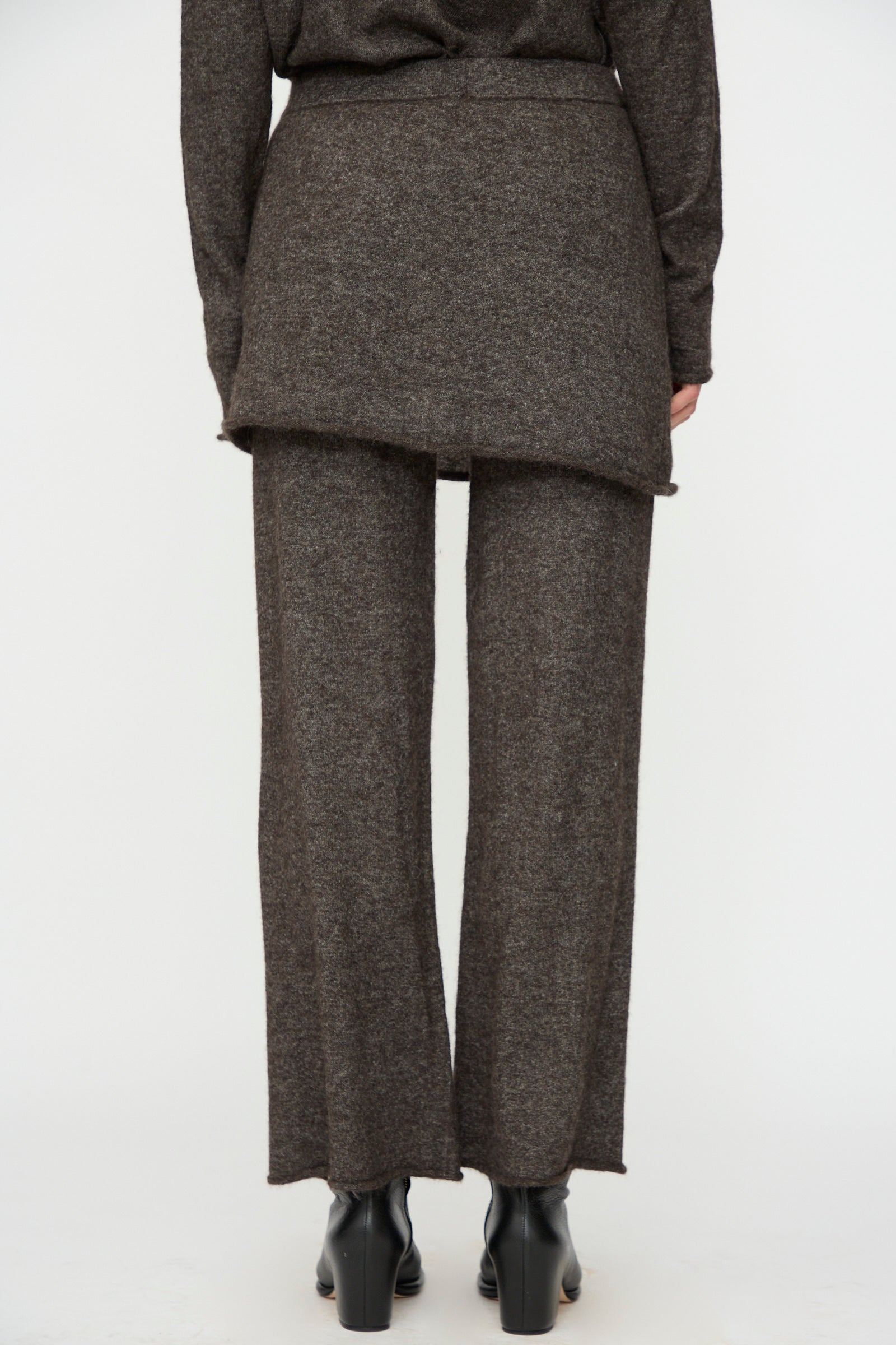 Person facing away, wearing Lauren Manoogian Askew Layer Pants in Blackened with a matching long top and black heeled shoes, standing against a plain light background.