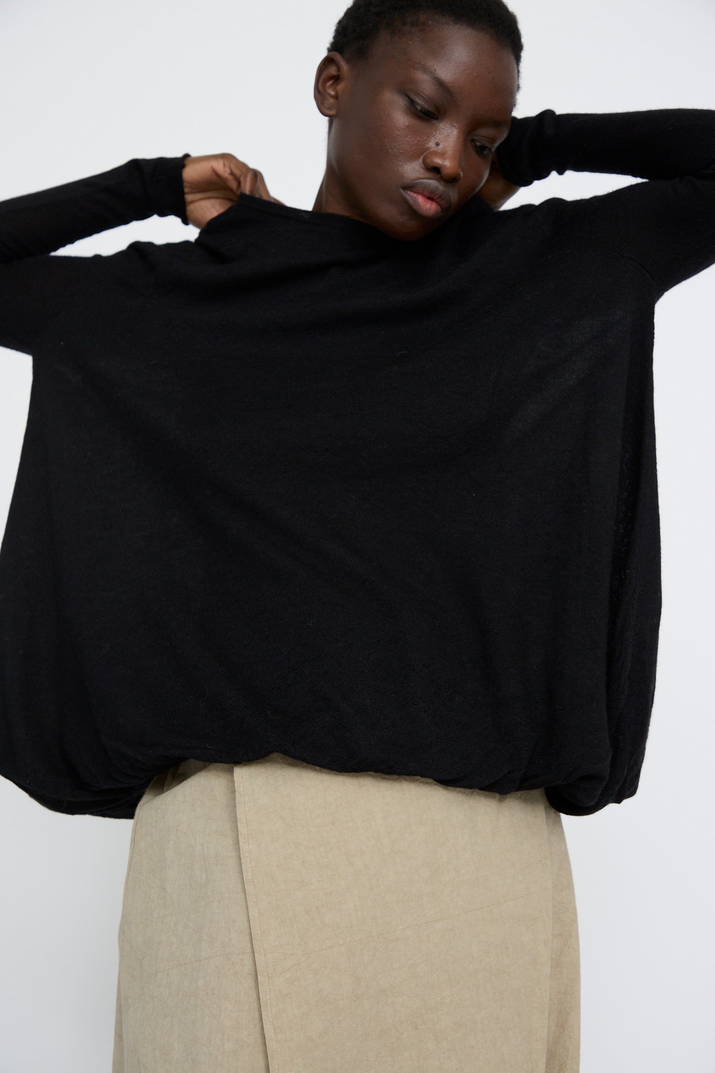 A person in an oversized Balloon Pullover in Black by Lauren Manoogian adjusts the collar over a beige skirt against a plain background.