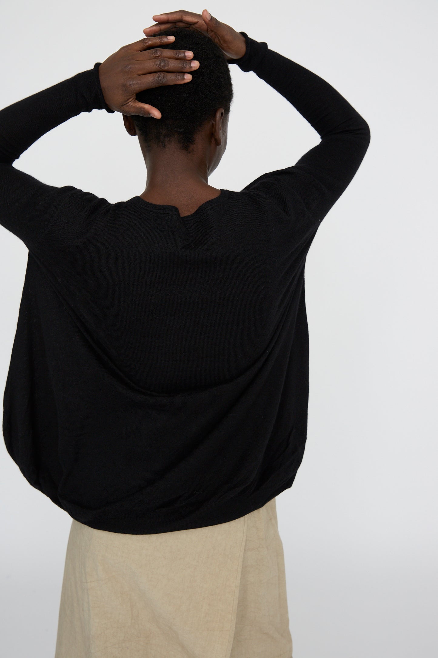 Facing away, a person wears the Balloon Pullover in Black by Lauren Manoogian, with an oversized fit and paired with a beige skirt. Their hands rest on the back of their head against a plain background.