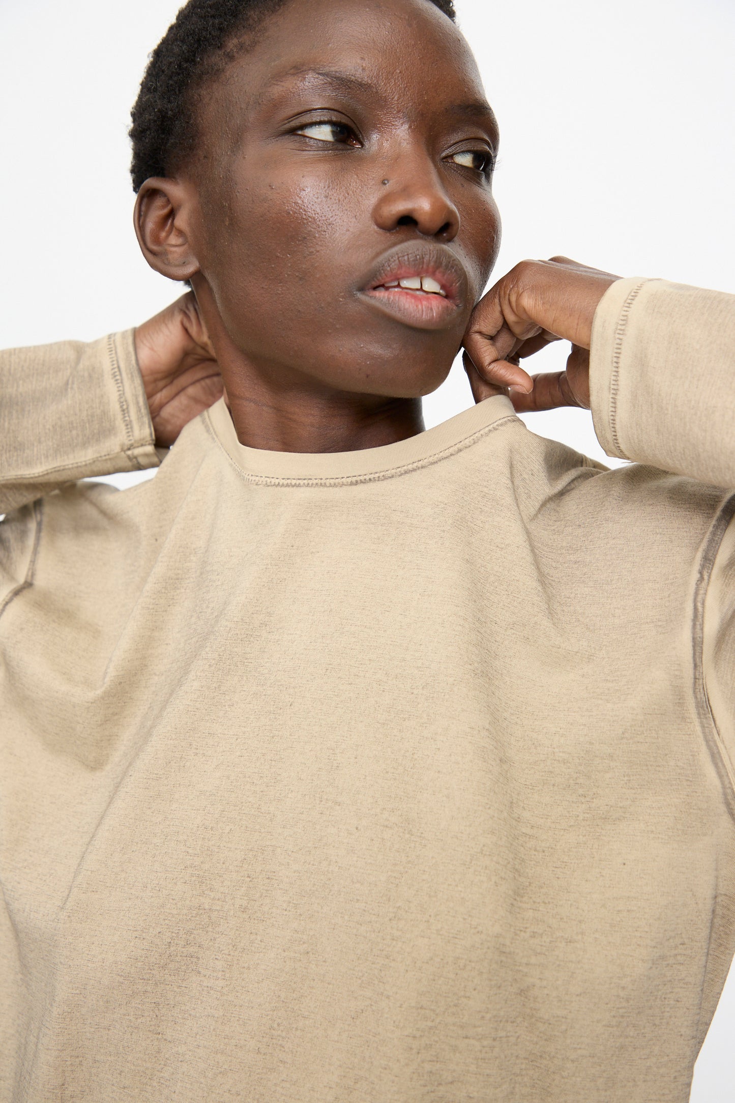 Person in the Lauren Manoogian Bias Long Sleeve Tee in Painted Antique adjusts their collar, looking off to the side against a plain light background.