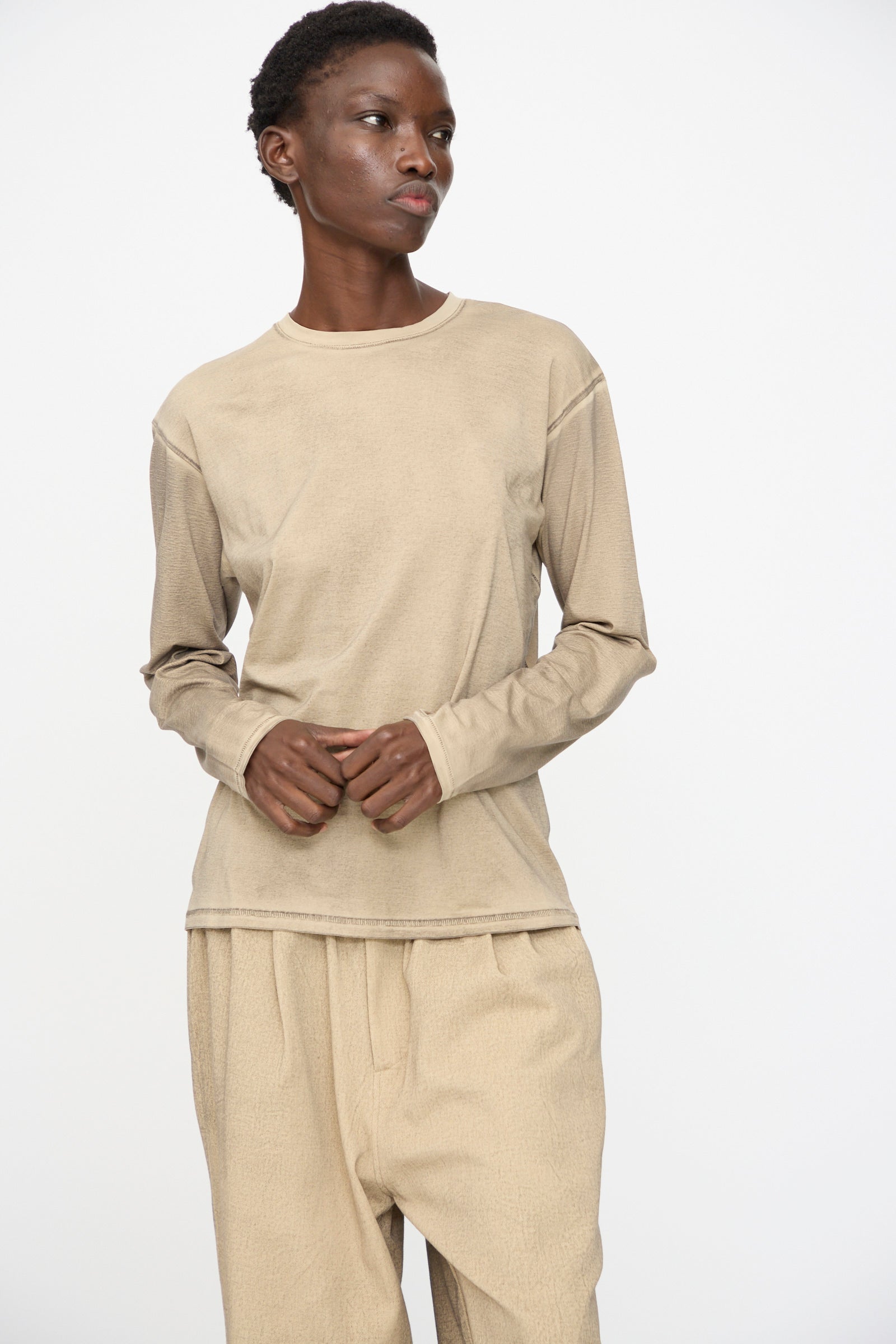 Person standing against a plain background, wearing the Lauren Manoogian Bias Long Sleeve Tee in Painted Antique and loose beige pants, looking slightly to the side with hands clasped.