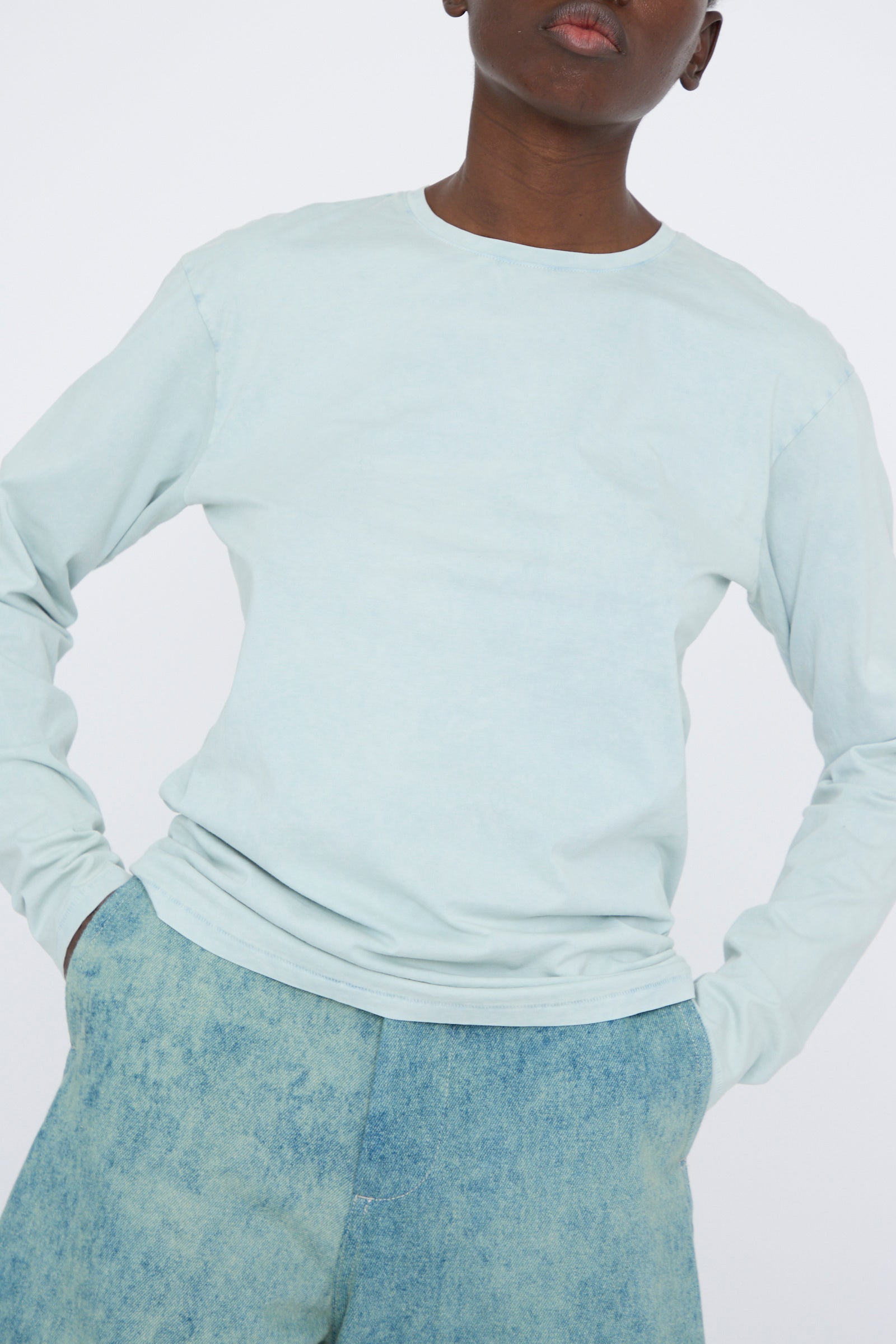 A person is wearing a relaxed fit, light blue Bias Long Sleeve Tee in Acid by Lauren Manoogian and blue pants, with hands in pockets against a plain background.