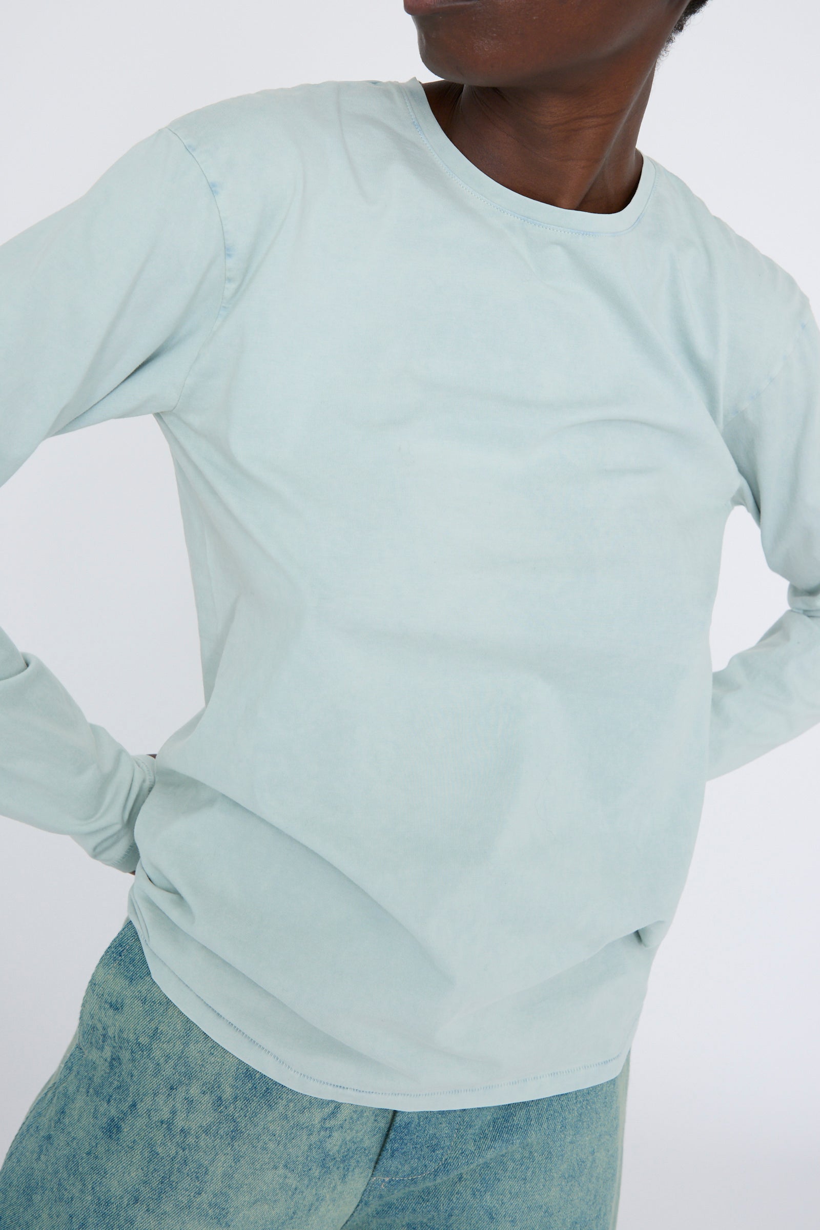 A person in a relaxed fit light blue Lauren Manoogian Bias Long Sleeve Tee in Acid and faded jeans stands with their hands on their hips against a plain white background.