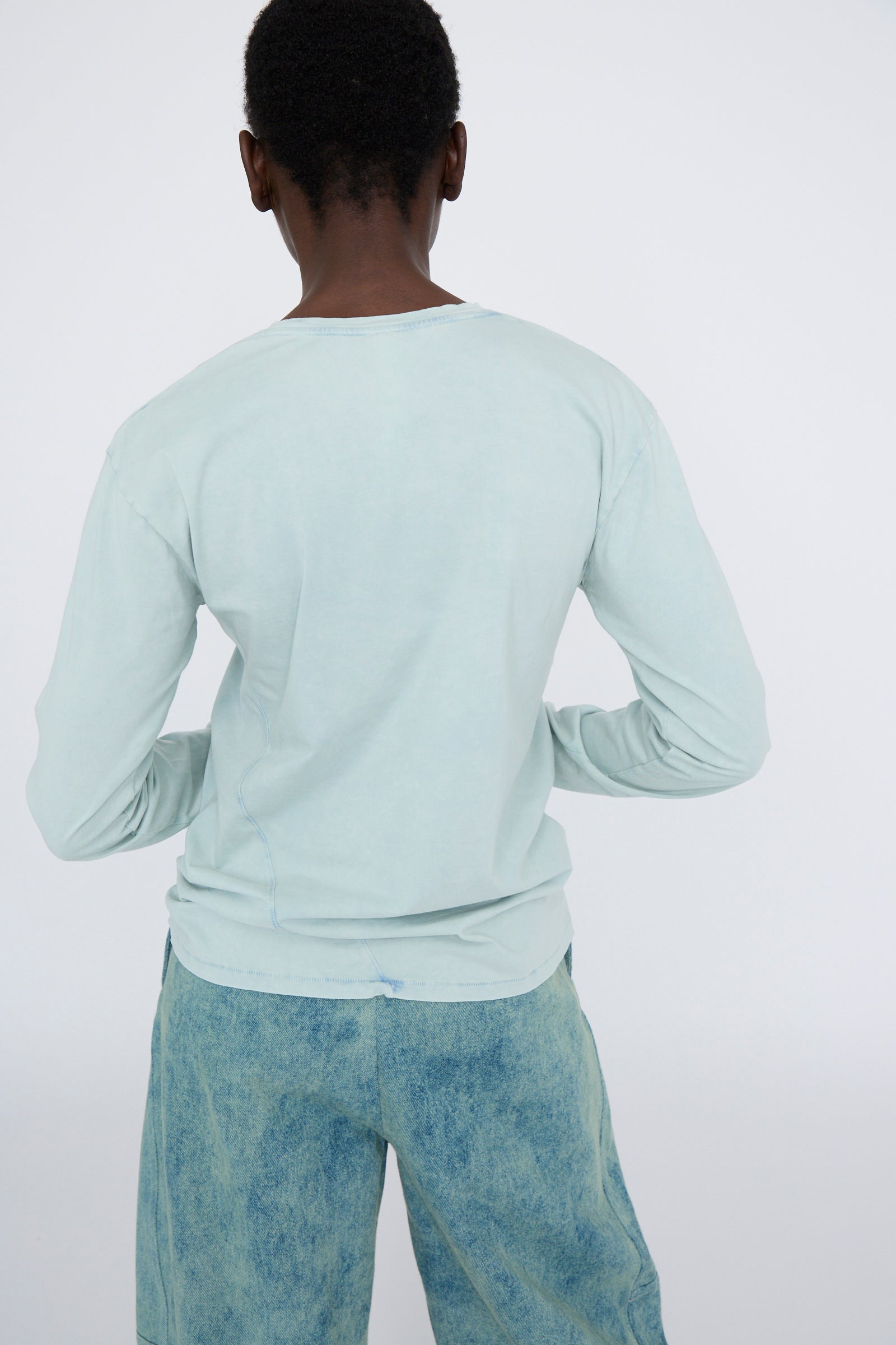 A person wearing the Lauren Manoogian Bias Long Sleeve Tee in Acid and faded blue jeans stands facing away from the camera against a plain background.