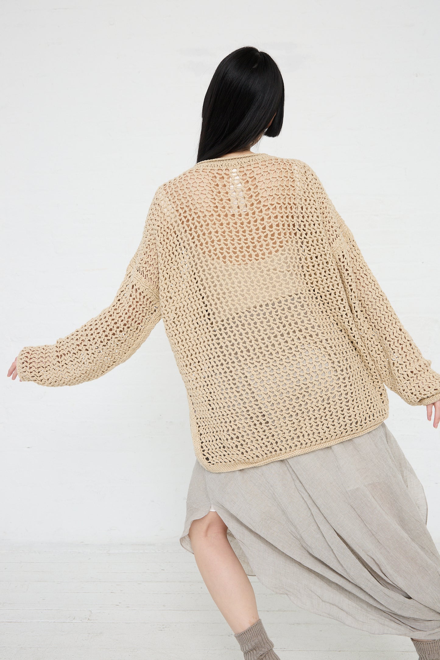 Woman posing with her back to the camera, wearing a beige oversized Lauren Manoogian Big Net Pullover in Jute and a flowing skirt made from sustainable knitwear.