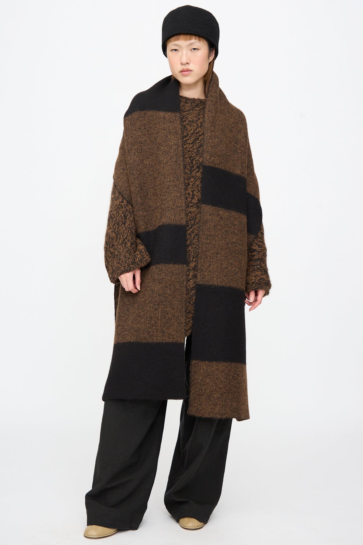 A person wears a long, Block Scarf in Rust Marl and Black draped over a matching sweater, with a black hat and wide-leg trousers.