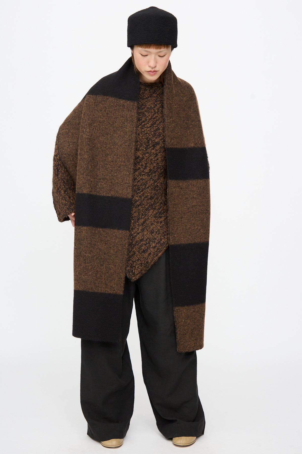 A person wears a Lauren Manoogian Block Scarf in Rust Marl and Black draped over a matching textured sweater, black trousers, and a black hat.
