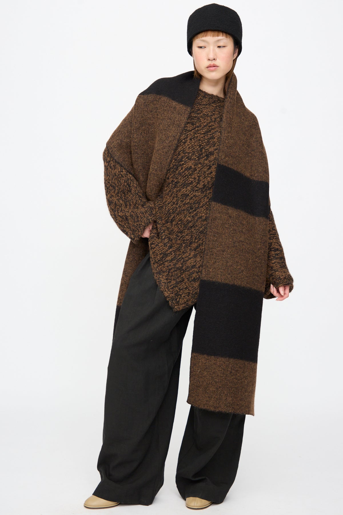 A person wearing a knit hat and a Lauren Manoogian Block Scarf in Rust Marl and Black, a long merino wool scarf featuring wide brown and black stripes.