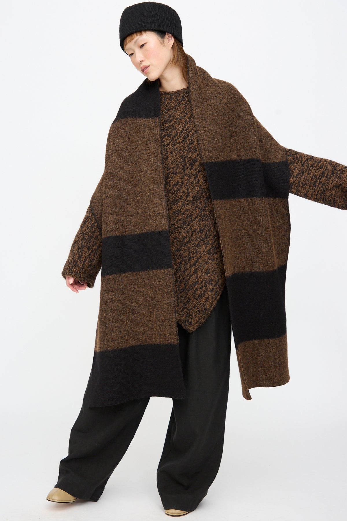 A model wears a Lauren Manoogian Block Scarf in Rust Marl and Black, a long, colorblocked merino wool scarf with wide brown and black horizontal stripes, over a matching sweater and black trousers.