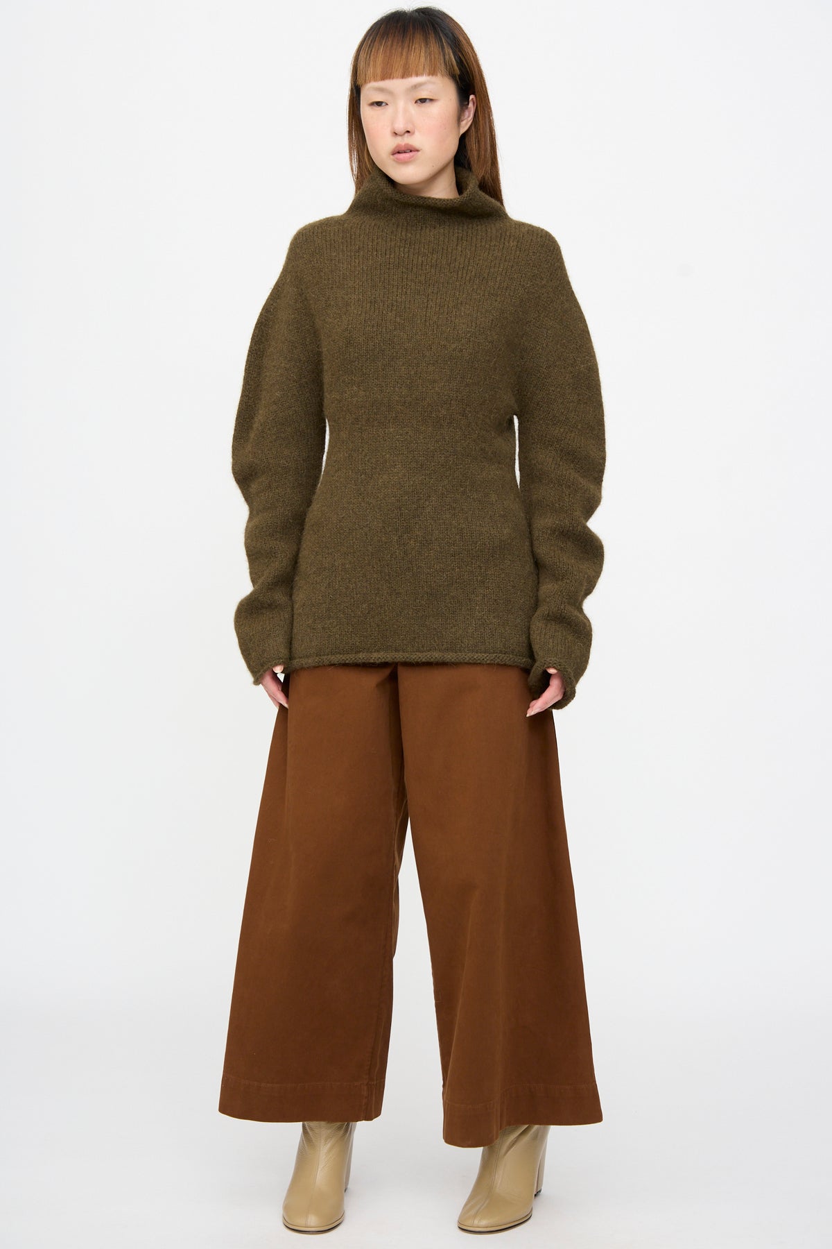 A person wearing a Lauren Manoogian Bottle Turtleneck in Olive, brown wide-leg trousers, and beige ankle boots.