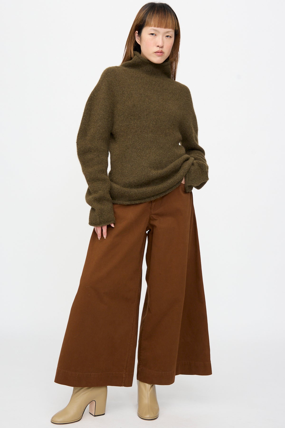 A model wearing a Lauren Manoogian Bottle Turtleneck in Olive, wide-leg brown trousers, and beige boots.