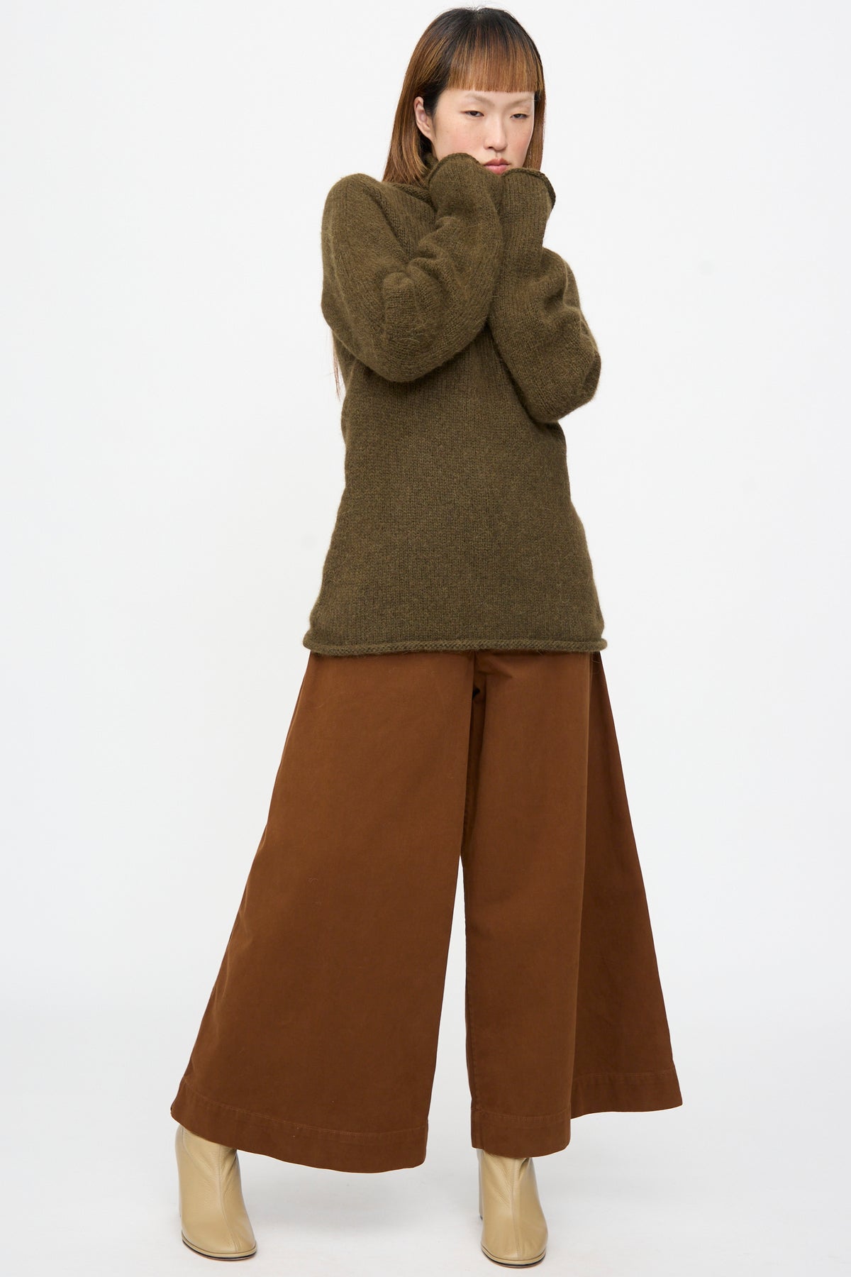 A person wearing a Lauren Manoogian Bottle Turtleneck in Olive and wide-leg brown trousers, with beige ankle boots.