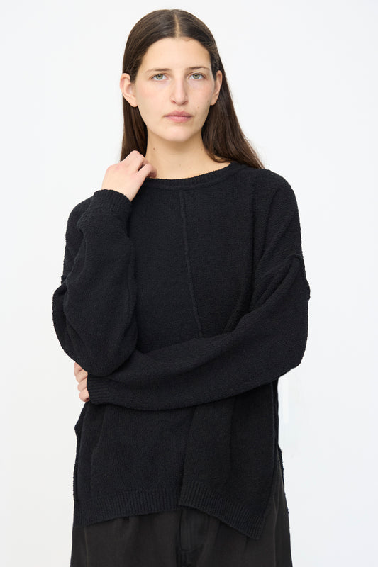 A person with long dark hair stands against a white background, wearing the Lauren Manoogian Boucle Crewneck in Black and black pants, with one hand touching their neck.