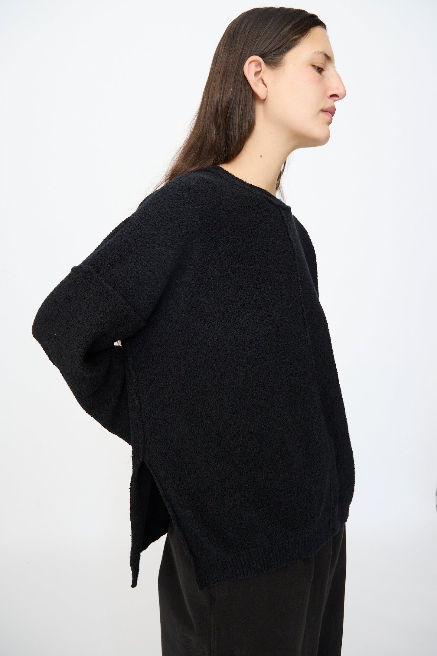 A person with long dark hair wears the Lauren Manoogian Boucle Crewneck in Black and black pants, standing in profile against a plain light background.