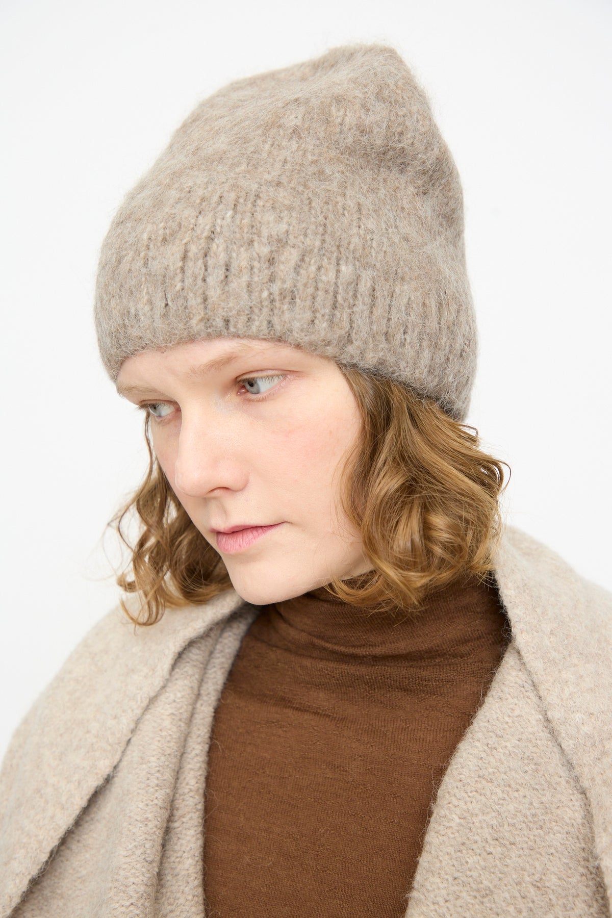 A person with light brown wavy hair wears a Lauren Manoogian Brushed Beanie in Grey Melange, paired with a brown turtleneck and beige cardigan, looking slightly downward against a plain light background.