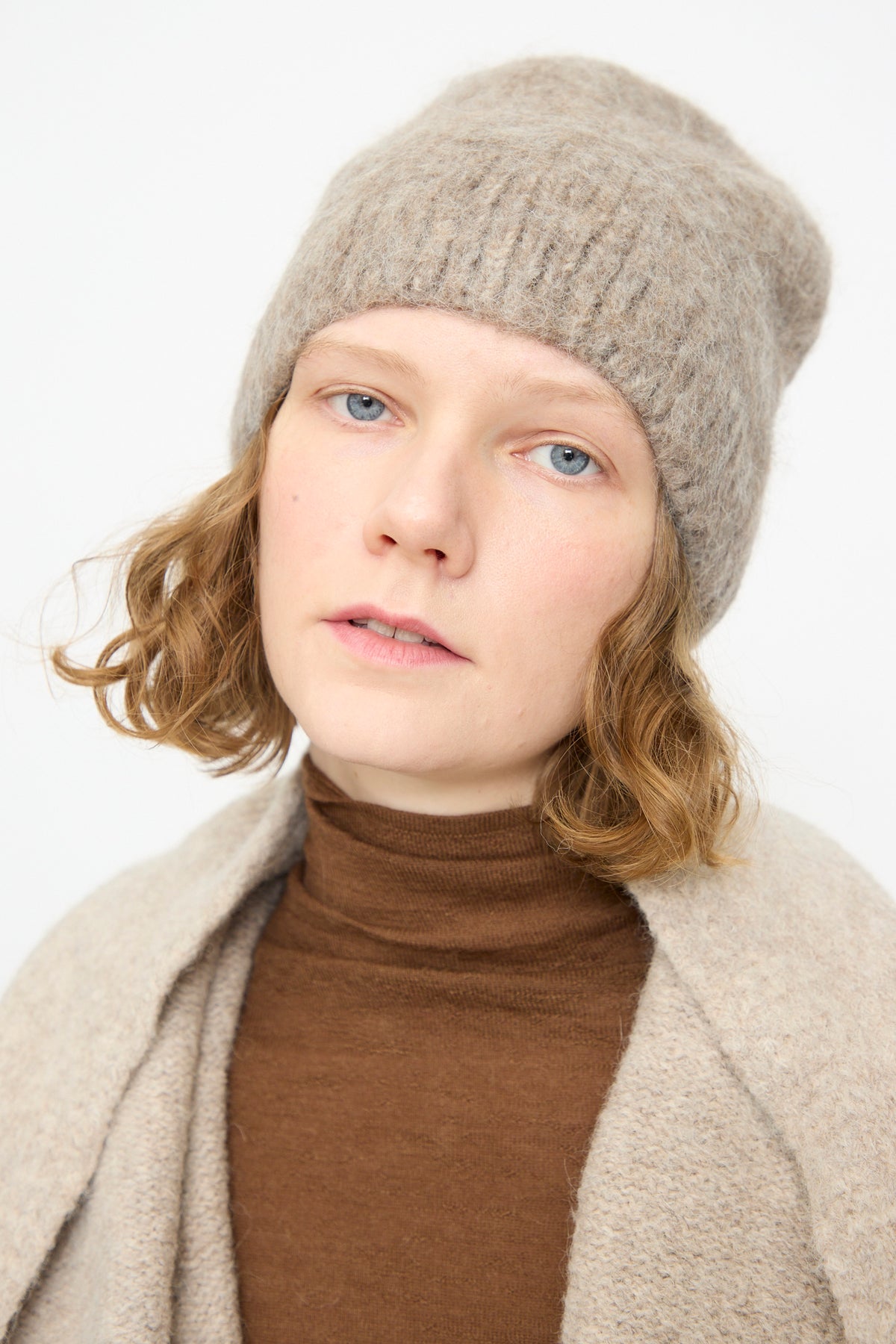 Person with light skin and wavy blonde hair wears the Lauren Manoogian Brushed Beanie in Grey Melange, a brown turtleneck, and a beige sweater, looking directly at the camera against a plain white background.