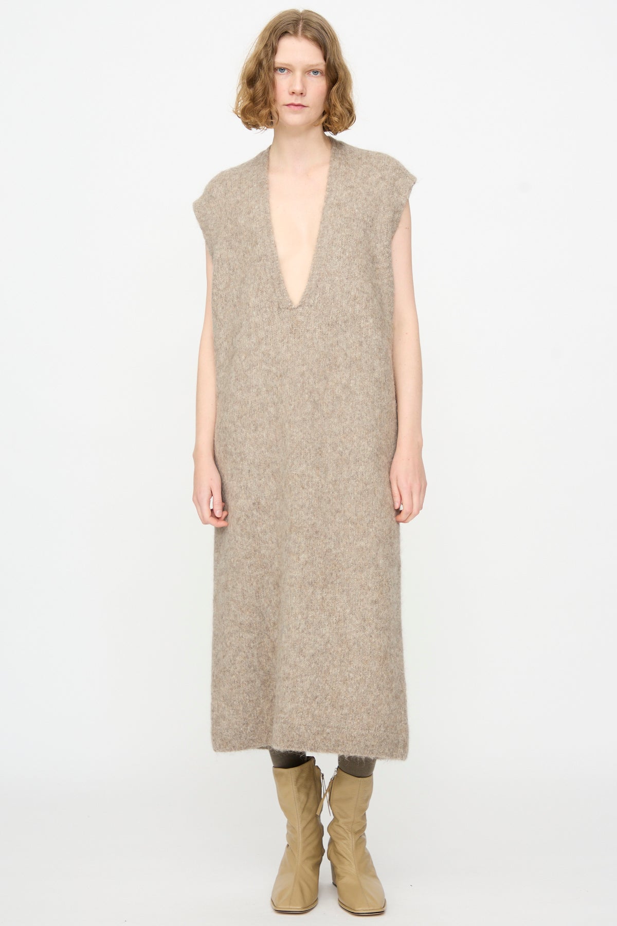 A person stands against a plain background wearing the Lauren Manoogian Brushed Low V Dress in Grey Melange—a luxurious, deep V-neck, sleeveless maxi dress crafted from a soft Suri alpaca blend and made in Peru—paired with light tan boots.