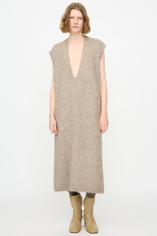 A person stands against a plain background wearing the Lauren Manoogian Brushed Low V Dress in Grey Melange—a luxurious, deep V-neck, sleeveless maxi dress crafted from a soft Suri alpaca blend and made in Peru—paired with light tan boots.