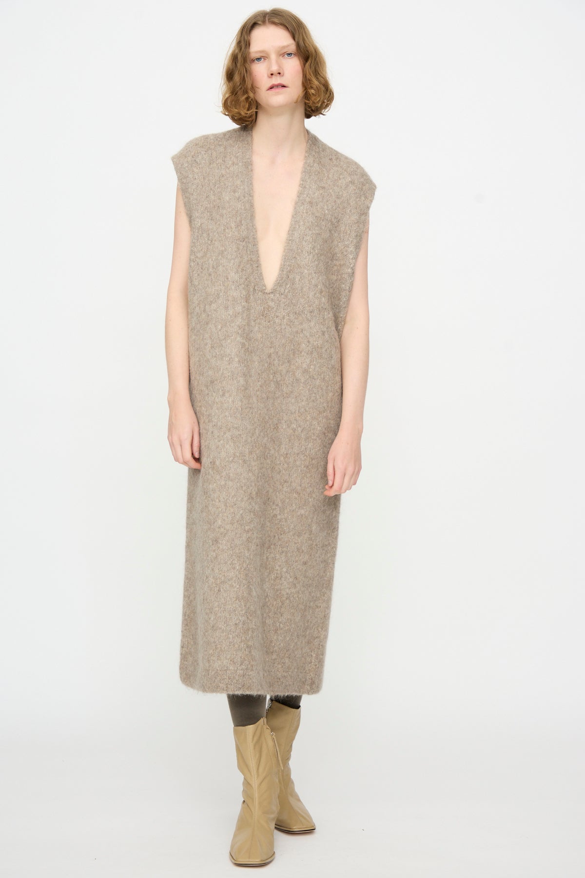 A person is standing against a plain background, wearing the Lauren Manoogian Brushed Low V Dress in Grey Melange—a maxi sleeveless dress with a deep V-neck, crafted from a luxurious Suri alpaca blend made in Peru—paired with gray tights and beige boots.