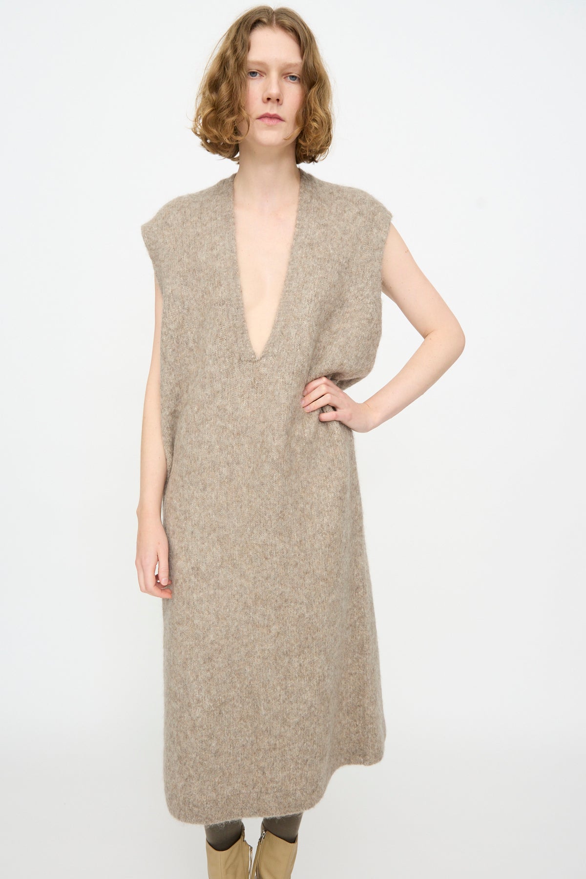 A person standing in a simple setting, wearing the Lauren Manoogian Brushed Low V Dress in Grey Melange—a sleeveless, knee-length knit dress crafted from a luxurious Suri alpaca blend with a deep V-neckline—paired with neutral boots.