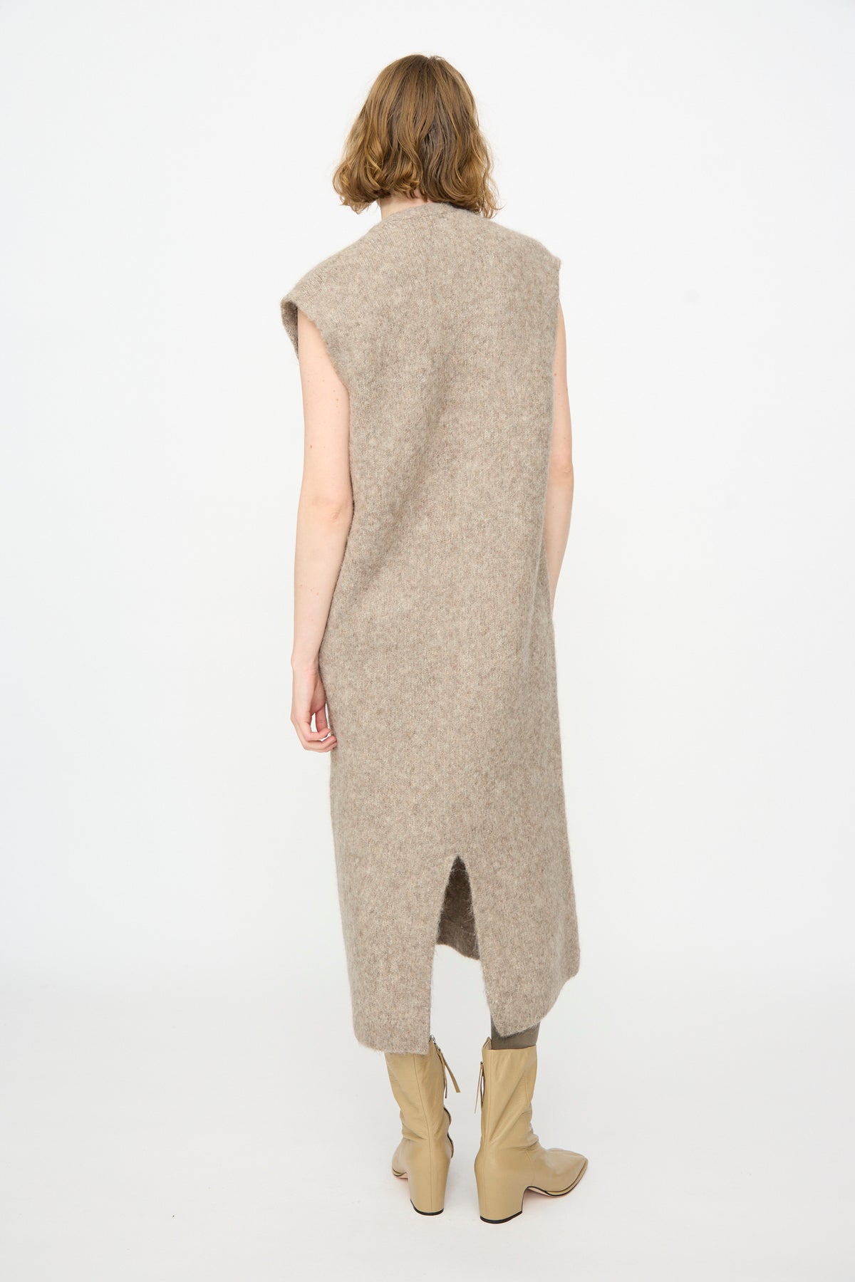 A person stands with their back to the camera, wearing the Lauren Manoogian Brushed Low V Dress in Grey Melange—a sleeveless, knee-length knit dress—paired with beige knee-high boots against a plain white background.
