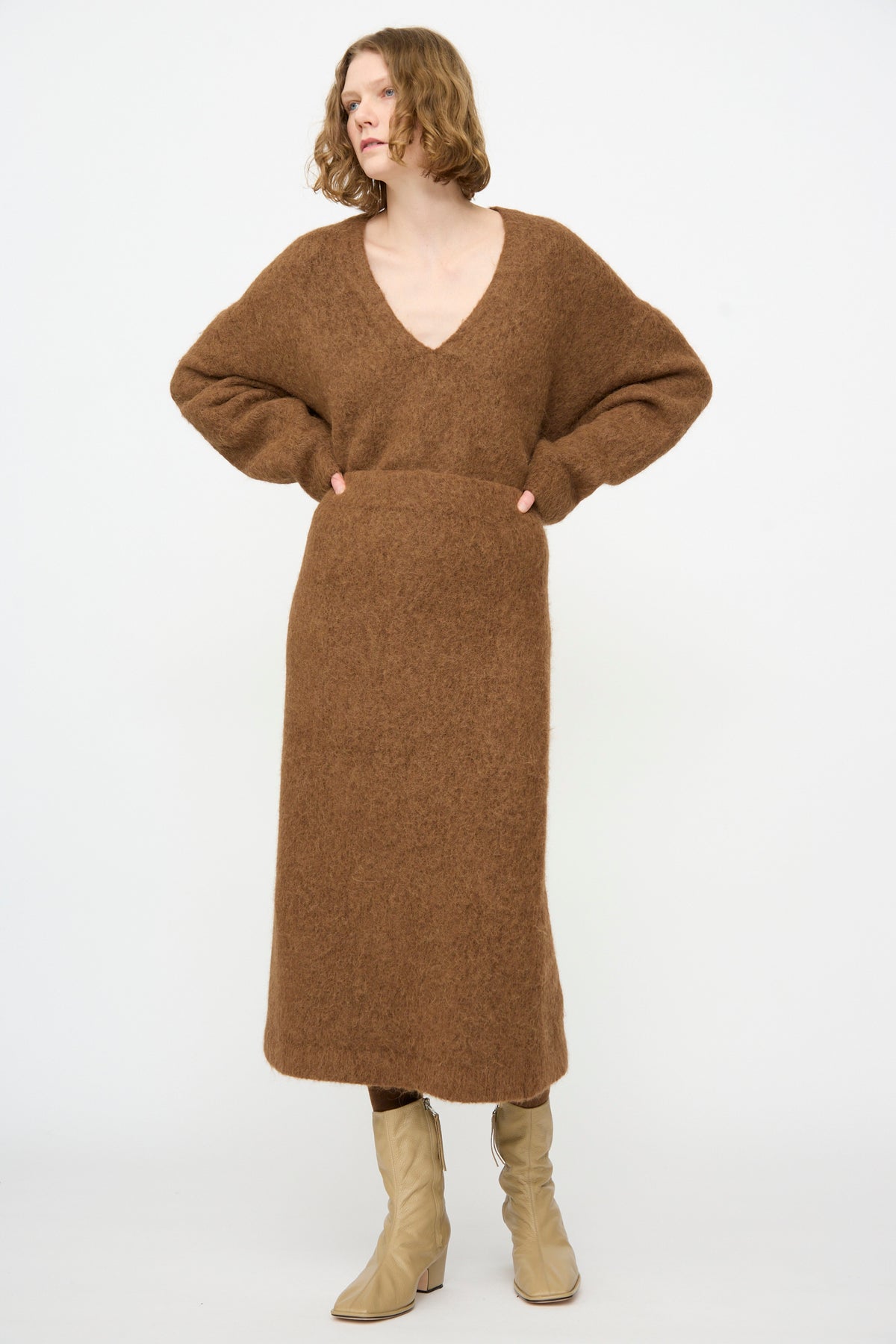 A person wearing the Lauren Manoogian Brushed Pencil Skirt in Rust, styled with a matching long-sleeved V-neck brown sweater and beige boots, stands against a plain white background.