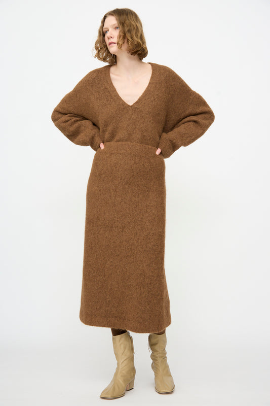 A person wearing the Lauren Manoogian Brushed Pencil Skirt in Rust, styled with a matching long-sleeved V-neck brown sweater and beige boots, stands against a plain white background.