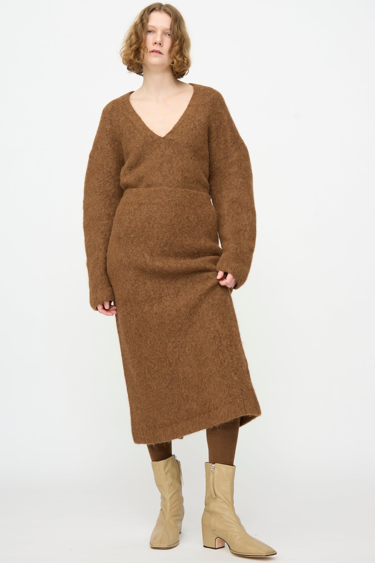 A person wearing a brown fuzzy long-sleeve dress with a V-neck, paired with the Lauren Manoogian Brushed Pencil Skirt in Rust—a cozy alpaca wool, chic midi-length skirt—along with brown leggings and beige ankle boots, stands against a plain white background.