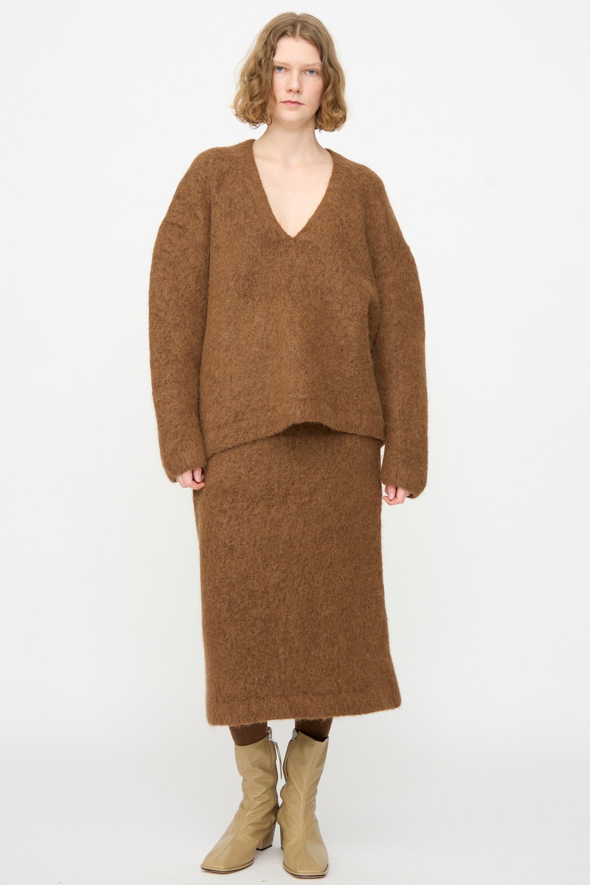 A person stands against a plain background wearing a loose brown V-neck sweater, the Lauren Manoogian Brushed Pencil Skirt in Rust, and beige boots.