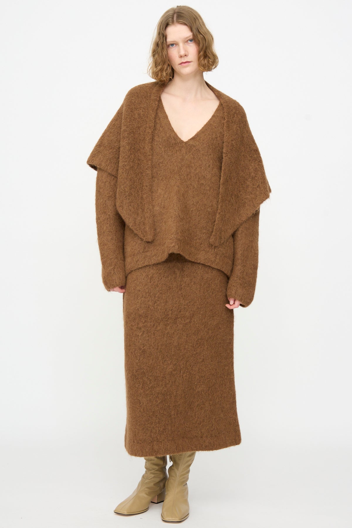 Wearing a brown fuzzy V-neck sweater, matching long skirt, and beige boots, the person stands against a plain white background, elegantly draping the Lauren Manoogian Brushed Triangle Wrap in Rust—handloomed in Peru—over their shoulders.