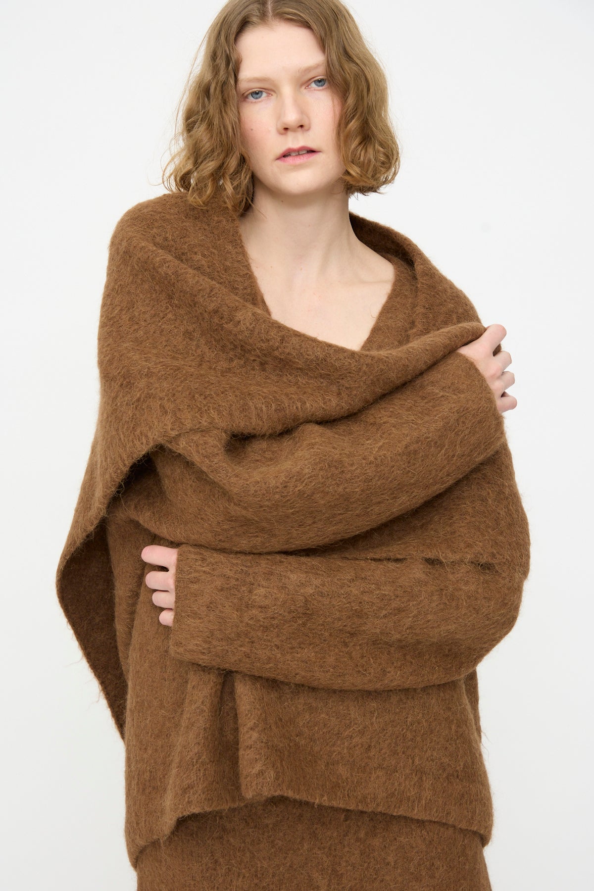 A person with wavy, light brown hair wears a loose brown fuzzy sweater and drapes the Lauren Manoogian Brushed Triangle Wrap in Rust around their shoulders, standing against a plain white background.