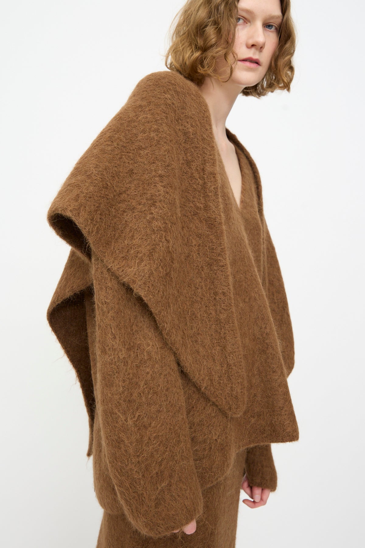 A person with short, wavy hair wears an oversized, textured brown sweater and matching pants against a plain white background, effortlessly accessorized with the elegant Brushed Triangle Wrap in Rust by Lauren Manoogian.