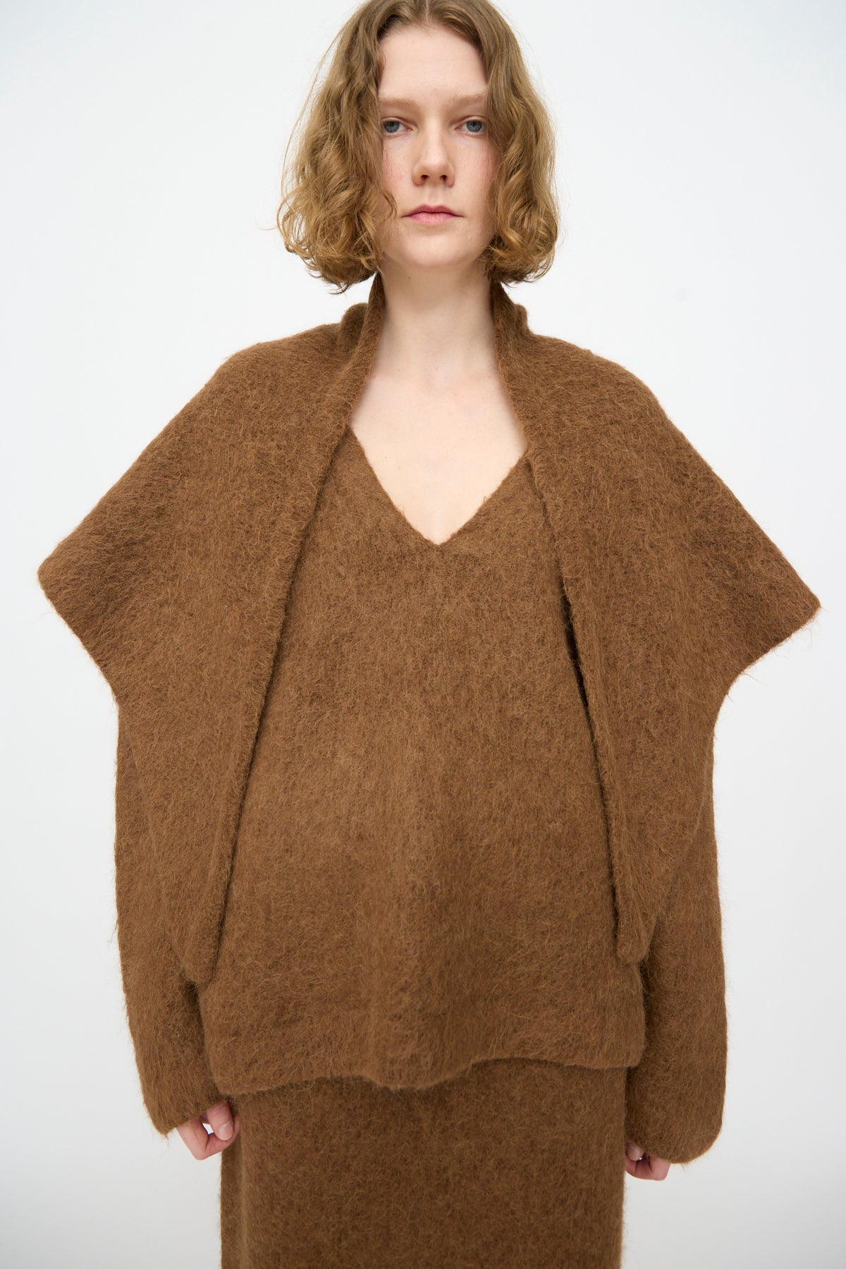 A person with short, wavy blonde hair wears a loose, textured brown sweater and matching skirt, standing against a plain white background, effortlessly draped in the Lauren Manoogian Brushed Triangle Wrap in Rust—handloomed in Peru.