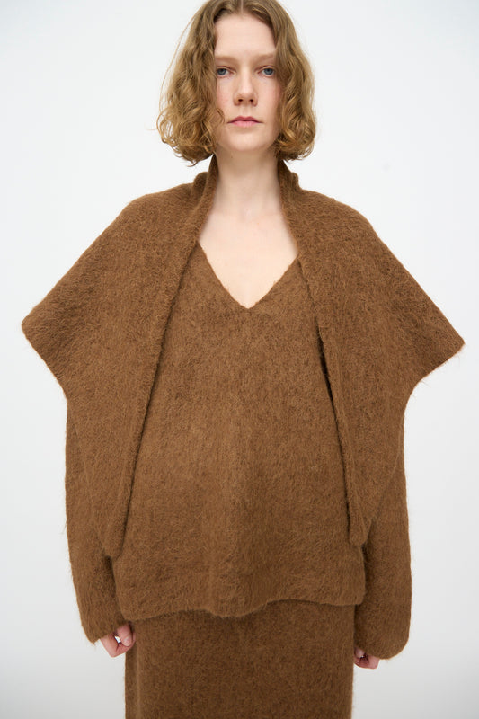 A person with short, wavy blonde hair wears a loose, textured brown sweater and matching skirt, standing against a plain white background, effortlessly draped in the Lauren Manoogian Brushed Triangle Wrap in Rust—handloomed in Peru.