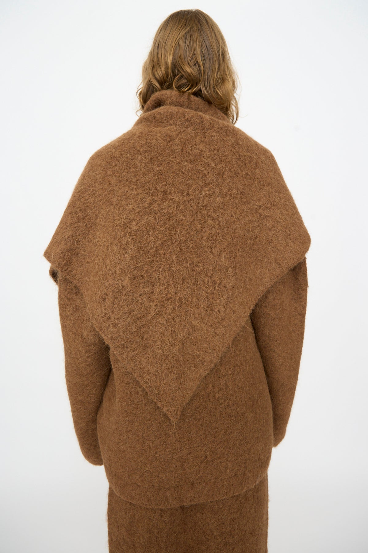 A person with shoulder-length wavy hair is photographed from the back against a plain white background, wearing the Lauren Manoogian Brushed Triangle Wrap in Rust—an artfully handloomed accessory draped over a matching brown sweater.