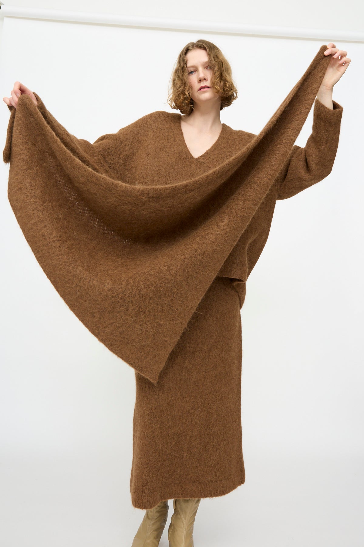 A person wearing the Lauren Manoogian Brushed Triangle Wrap in Rust holds out one side of the wrap sweater, standing against a plain white background.