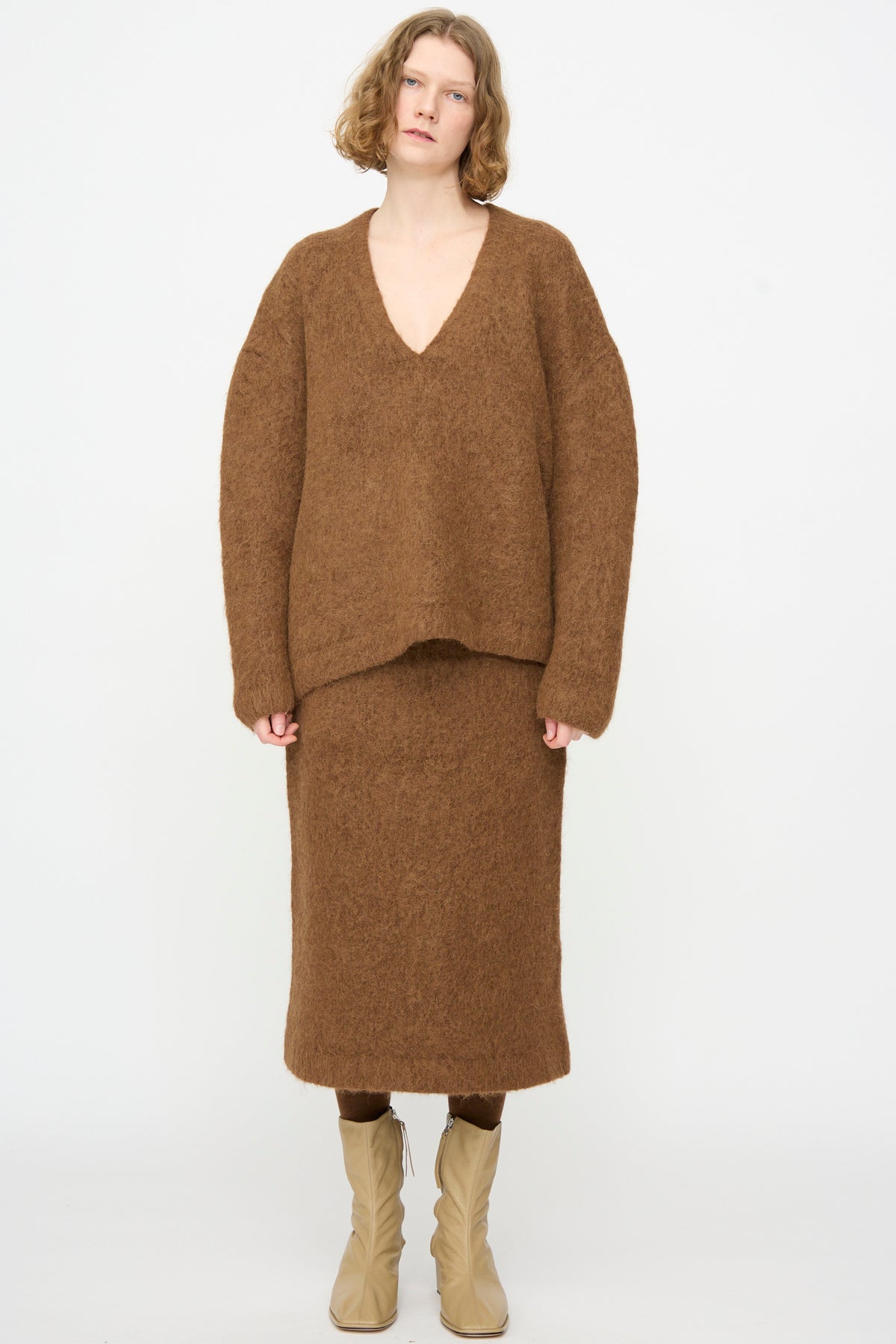 Person stands facing forward, wearing an oversized fit Lauren Manoogian Brushed V Neck in Rust sweater and matching midi skirt with tan ankle boots, against a plain white background.