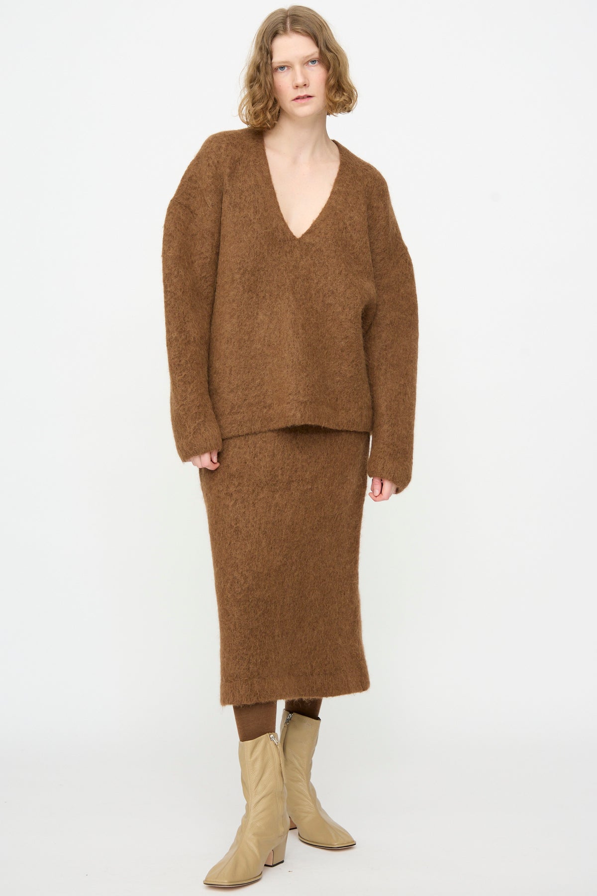 A person is standing against a white background wearing the Lauren Manoogian Brushed V Neck in Rust—an oversized fit Suri Alpaca sweater—paired with a matching brown skirt and beige ankle boots.